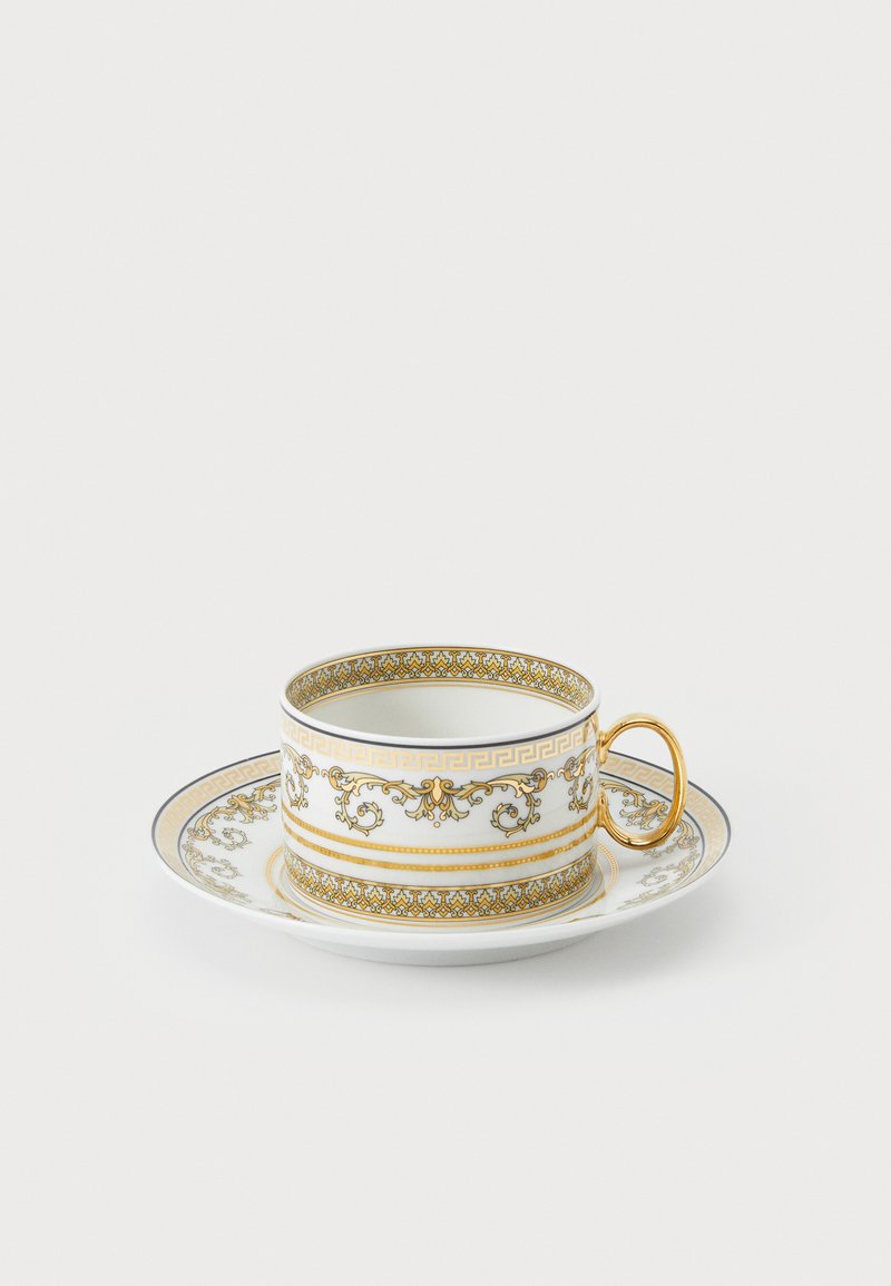 Taza y plato de porcelana con detalles dorados que presentan patrones intrincados, fondo blanco y un asa decorativa. Textura suave y brillante.