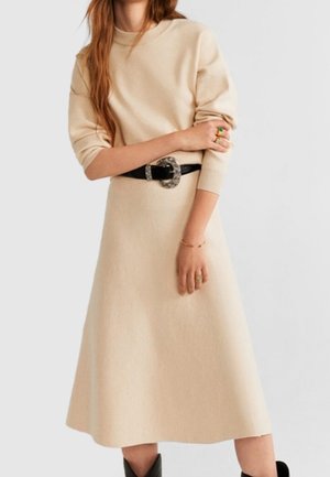 Woman wearing beige long-sleeve dress with black belt and decorative buckle, standing with arms crossed, wearing rings and bracelet.