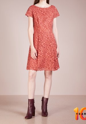 Vrouw in een kniehoog rode kanten jurk met korte mouwen en bordeauxrode enkellaarsjes, staand op een beige vloer tegen een effen achtergrond.