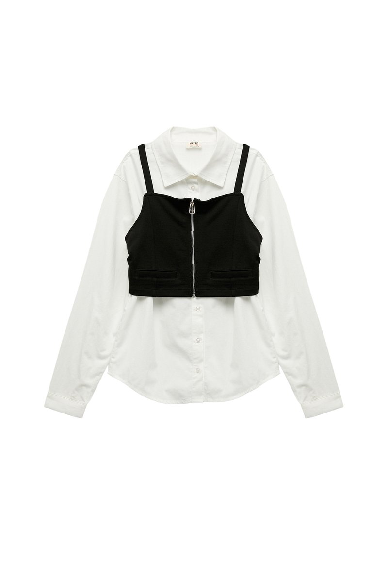Top corto nero con bretelle regolabili sopra una camicia bianca a maniche lunghe, caratterizzato da una cerniera anteriore e due piccole tasche frontali.