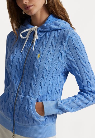 Lichtblauwe kabelgebreide zip-up hoodie met capuchon, witte trekkoord, geribde manchetten en een klein geel logo op de borst.