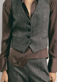 Grahameliertes Wollweste mit schwarzen Knöpfen über einem braunen Seidenhemd. Strukturierter Stoff und geschichtetes Design mit maßgeschneidertem Schnitt.