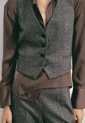 Chaleco de lana gris con botones negros sobre una camisa de seda marrón. Tejido texturizado y diseño en capas con un ajuste a medida.
