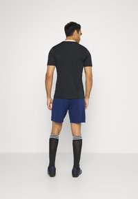 Camiseta negra de manga corta, pantalones cortos azules, calcetines negros hasta la rodilla con rayas blancas y zapatos de fútbol negros, vistos desde atrás.
