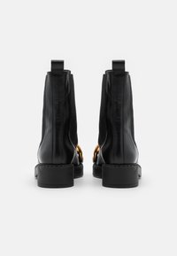 Paire de bottines en cuir noir avec talons blocs et détail chaîne dorée, vues de dos sur un fond blanc.