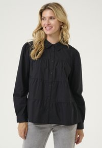 Camisa negra de botones con cuello, diseño en capas y mangas largas. Confeccionada en una tela suave y de corte relajado.