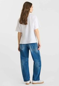 Femme aux longs cheveux bruns portant un T-shirt blanc ample, un jean bleu et des baskets blanches, se tenant de dos devant un fond uni.