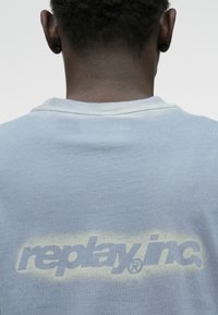 Ανοιχτό μπλε βαμβακερή φούτερ με στρογγυλή λαιμόκοψη, featuring ένα ξεθωριασμένο λευκό λογότυπο "replay.inc" εκτυπωμένο στην πλάτη. Απαλή υφή, casual σχέδιο.