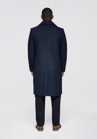 Marineblå uldfrakke med bred revers, lange ærmer og lige snit, kombineret med sorte bukser og brune sko.