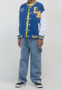 Niebiesko-biała kurtka varsity z żółtym napisem "LA", zestawiona z żółtą koszulką z grafiką i oversize’owymi niebieskimi dżinsami. Czarne trampki.