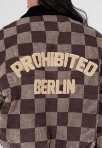 Femme portant une veste en velours côtelé marron et beige à carreaux avec "PROHIBITED BERLIN" brodé au dos en lettres crème.