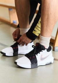 Scarpe sportive in bianco e nero con accenti rossi, caratterizzate da una rete testurizzata e sovrapposizione trapuntata, allacciate con lacci neri su una superficie liscia.