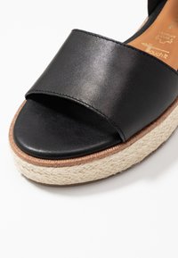 Svart läder sandal med öppen tå och flätad beige plattformssula samt tanfärgad innersula som visar varumärkespräglingar.