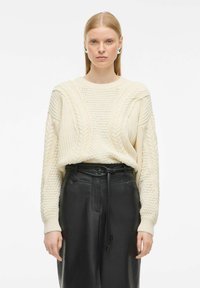 Pull en crème tricoté avec des motifs en relief, coupe ample et poignets côtelés, associé à un pantalon en cuir noir taille haute avec une ceinture.