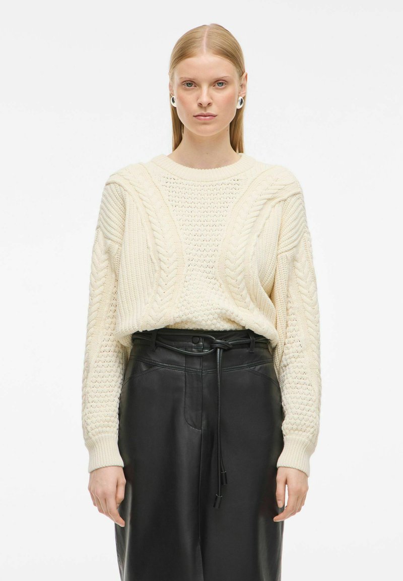 Pull en crème tricoté avec des motifs en relief, coupe ample et poignets côtelés, associé à un pantalon en cuir noir taille haute avec une ceinture.