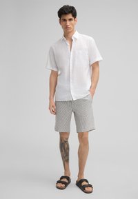 Weißes Kurzarmhemd mit Knöpfen und Tasche, kombiniert mit hell gestreiften Shorts. Schwarze Sandalen vervollständigen das Outfit. Glatte Textur.
