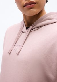 Νεαρό άτομο που φοράει ανοιχτό ροζ φούτερ Puma με κορδόνια και ανάγλυφο λογότυπο, που δείχνει το κάτω μέρος του προσώπου και το επάνω μέρος του στήθους.