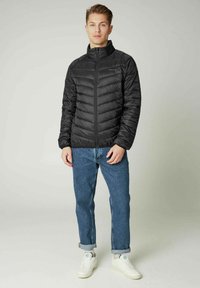 Sort polstret jakke med høj krave, med diagonale syninger og en frontlynlås, parret med blå denim jeans og hvide sneakers.