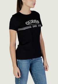 T-shirt noir en coton à manches courtes et col ras du cou ; présente le logo « GUESS » en argent avec des accents en strass, associé à un jean bleu.