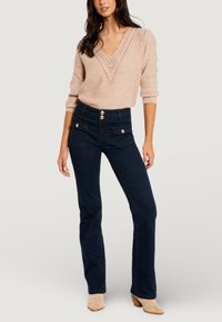 Suéter de punto rosa claro con cuello en V y diseño de punto abierto, combinado con jeans acampanados de talle alto en azul oscuro con botones dorados.
