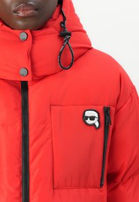 Red puffer jacket with a high collar, featuring a zipper and snap buttons; includes a patch on the chest pocket and a drawstring at the hood.