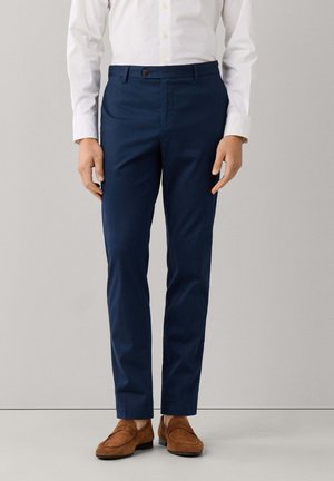 Man draagt marineblauwe slim-fit broek, wit overhemd met knopen en bruine suède loafers, staat tegen een effen achtergrond.