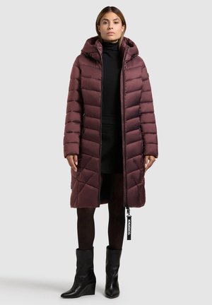 Doudoune bordeaux avec capuche, fermeture éclair, design matelassé, poches latérales et étiquette de la marque. Portée par-dessus un haut et une jupe noirs.