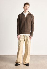 Barna texturált kötött pulóver széles gallérral, bézs bőszárú nadrággal és fekete cipővel, semleges háttér előtt állva.