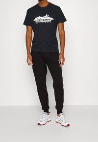 Musta lyhythihainen t-paita, jossa on vuoristografiikka ja valkoinen teksti, yhdistetty mustiin collegehousuihin ja valkoisiin lenkkareihin, joissa on punaisia yksityiskohtia.