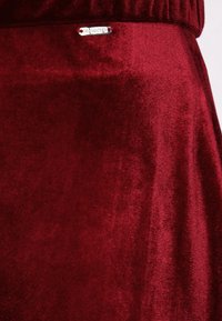 Burgundy samettihame, jossa on sileä pinta ja hopeinen logotagi, esittelee rikasta väriä ja pehmeää kiiltoa.