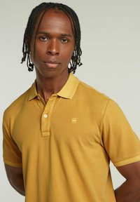 Polo shirt giallo senape in cotone, con colletto, patta con tre bottoni e un piccolo logo sul lato sinistro del petto. Texture liscia.