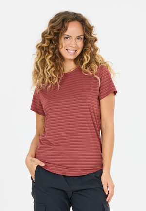 Lachende vrouw met krullend haar, gekleed in een roestrood gestreept shirt met korte mouwen en zwarte cargobroek, staand met één hand in haar zak.