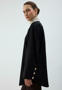 Maglia nera a maniche lunghe con un dolcevita alto color crema. Presenta dettagli con bottoni dorati sui polsini e una vestibilità morbida. Tessuto dalla texture liscia.