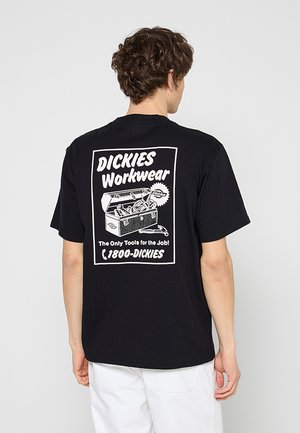 Hombre con camiseta de trabajo Dickies negra con gráfico blanco de una caja de herramientas y texto en la espalda, combinada con pantalones blancos, de espaldas.