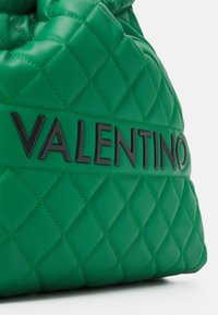 Grön quiltad läderhandväska med texturerad yta. Har en svart präglad "VALENTINO"-logotyp på ett slätt band.