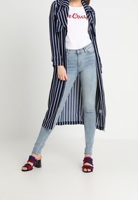 Cappotto lungo a righe navy e bianco sopra una t-shirt bianca con testo rosso, abbinato a jeans skinny blu chiaro e sandali con tacco colorati.