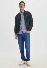 Marineblaues Bomberjackett mit gerippten Manschetten über einem hellblau-weiß gestreiften Hemd, kombiniert mit blauen Jeans und cremefarbenen Sneakern.