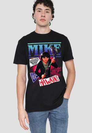 Junger Mann trägt ein schwarzes Stranger Things T-Shirt mit der Figur Mike, die einen Hockeyschläger hält, mit sichtbaren Hawkins- und Show-Logos.
