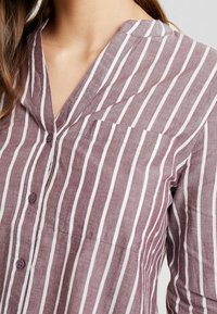 Chemise en lin avec rayures verticales bordeaux et blanches, col en V et poche poitrine. Texture lisse et fermeture boutonnée.