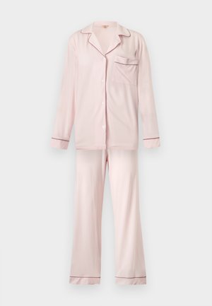 Femme debout pieds nus portant un haut de pyjama rose clair à manches longues boutonné et un pantalon ample assorti sur un fond blanc uni.