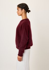 Femme aux cheveux tressés en chignon bas, portant des boucles d'oreilles créoles dorées, un pull bordeaux en tissu duveteux et une jupe gris clair, debout de profil.