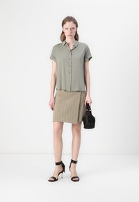 Light olive green short-sleeve blouse with button closure, paired with a beige pinstriped skirt. Black strappy sandals and a small black bag.
