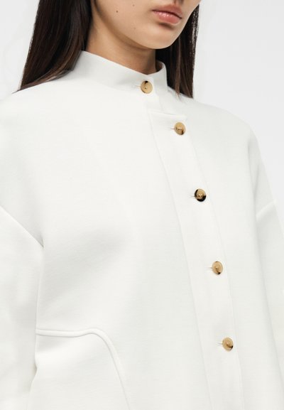 Λευκό μπουφάν με κουμπιά, με ψηλό γιακά, ξύλινα κουμπιά και λεία υφή. Διαθέτει πλαϊνές τσέπες και μινιμαλιστικό σχέδιο.