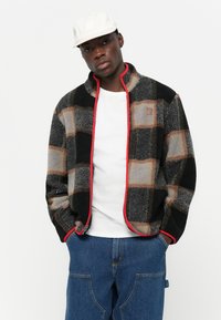 Mikina z fleeceu s čiernym, šedým a béžovým kockovaným vzorom s červenými akcentmi a so zipsom vpredu. Nosená cez biele tričko a modré džínsy.