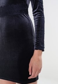 Mörkblå sammetsklänning med långa ärmar, med en spetspanel vid midjan och fint texturerade ärmar. Slät tygfinish.