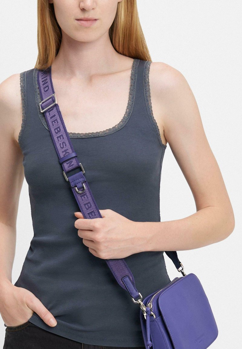 Femme portant un débardeur gris foncé avec une bordure en dentelle, portant un sac bandoulière violet avec une large sangle réglable sur l'épaule.