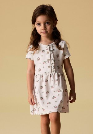 Ung jente med langt brunt hår har på seg en hvit kortermet kjole med små blomster- og jordbærmønstre, står mot en beige bakgrunn.