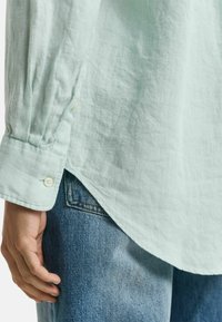 Mintgroene linnen shirt met een ontspannen pasvorm, voorzien van een knoopmanchet en een gebogen zoom, gecombineerd met lichtblauwe denim jeans.