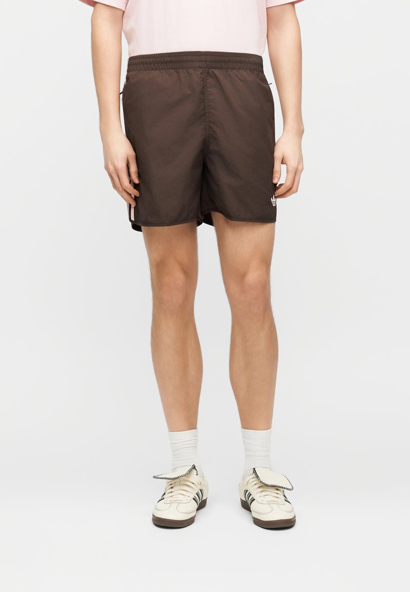 Persona che indossa pantaloncini sportivi marrone scuro, calzini bianchi alla caviglia e sneaker crema con dettagli neri, in piedi contro uno sfondo semplice.