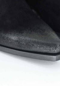 Primo piano di una scarpa nera a punta con superficie texturizzata e suola piatta su uno sfondo bianco.